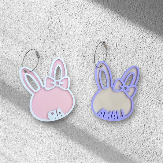 Two bunny-shaped earrings, one pink and one purple, on a light gray background.